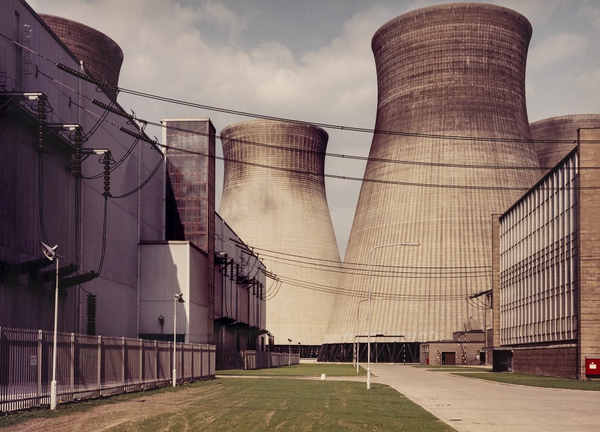 Photograph of power station courtyard with two large chimneys in the background, substation on the left-hand side and building on the right, with electric cables connecting the two buildings across the yard