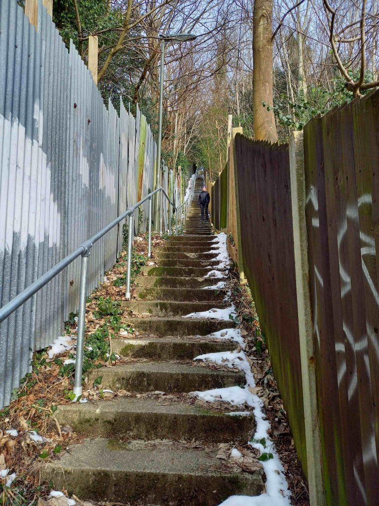 A steep flight of concrete steps and a woman standing in the middle far distance. There is a high corrugated metal fence on the left hand side and a wooden slatted fence on the right hand side. There is snow on the edges of the steps.
