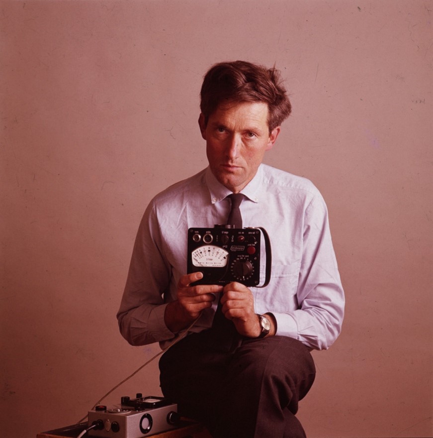 A colour photograph of Bryan Heseltine, a white man in a shirt and tie, sitting and holding an electronic device.