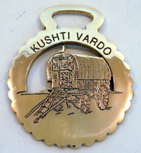 This horse brass is round with scalloped edges. It is engraved with a picture of a caravan and the inscription 'Kushtie Vardo', which means 'A Fine Wagon'. The caravan depicted is a Bill Wright Bow Top Wagon (Leeds from c. 1908). The back of the brass is stamped 'KB 61'.